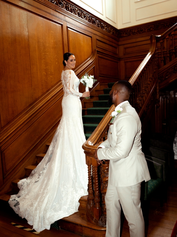 Bride In A Lace Detail White Gown Standing On A Grand Wooden Staircase As The Groom Looks Up At Her From The Base.
